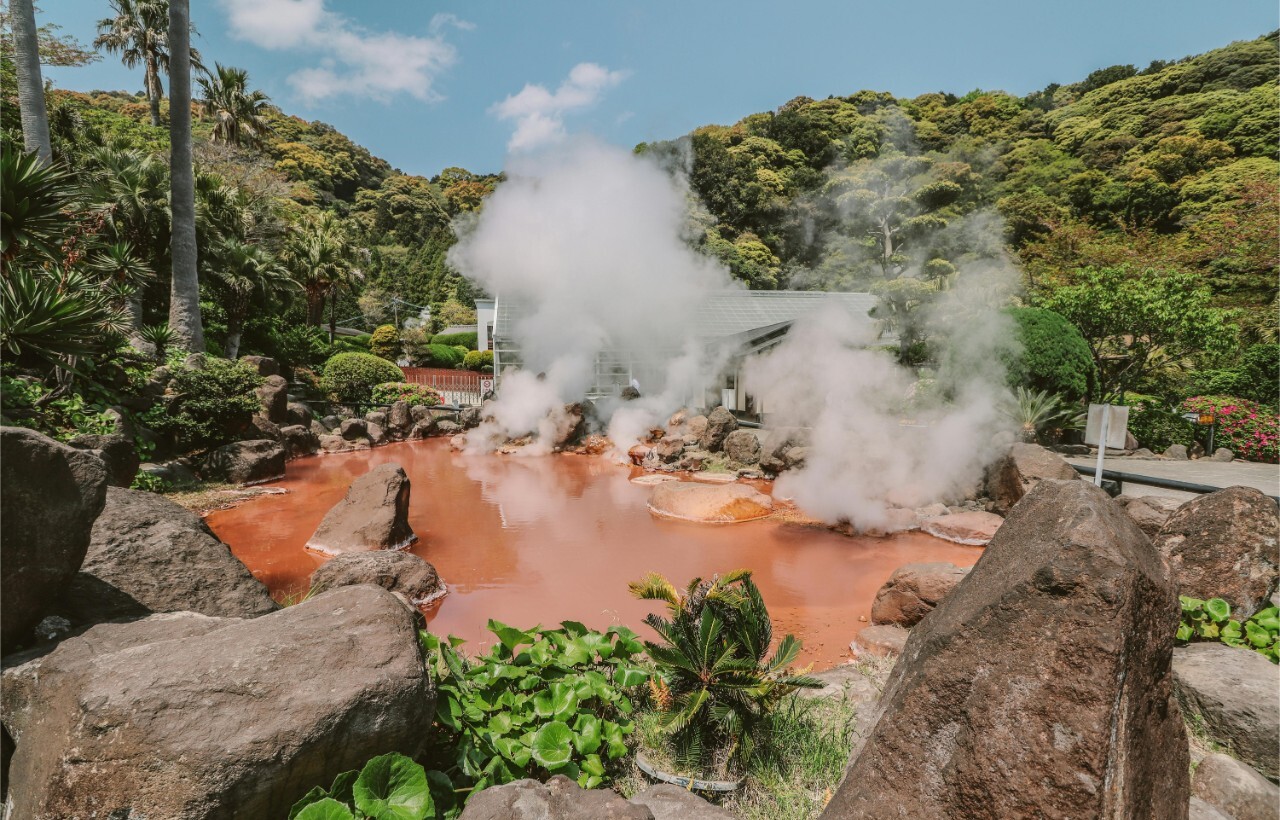 A steaming, red-hued hot spring is encircled by rocks and lush greenery beneath a clear sky.