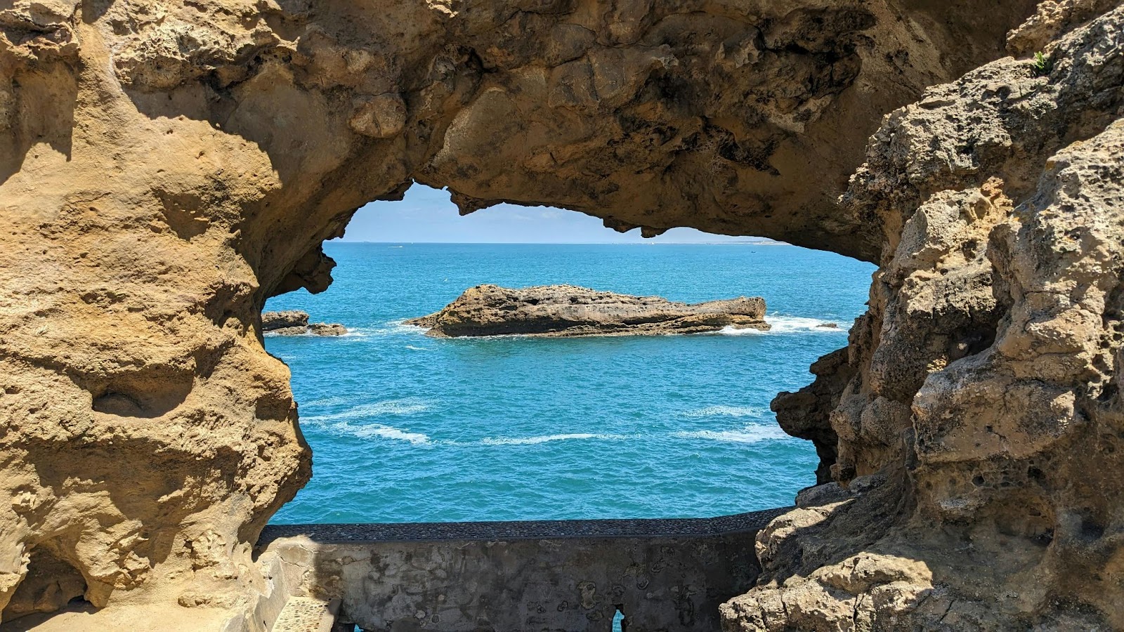 A natural rock formation frames an ocean view with a large rock in the water and a clear blue sky.