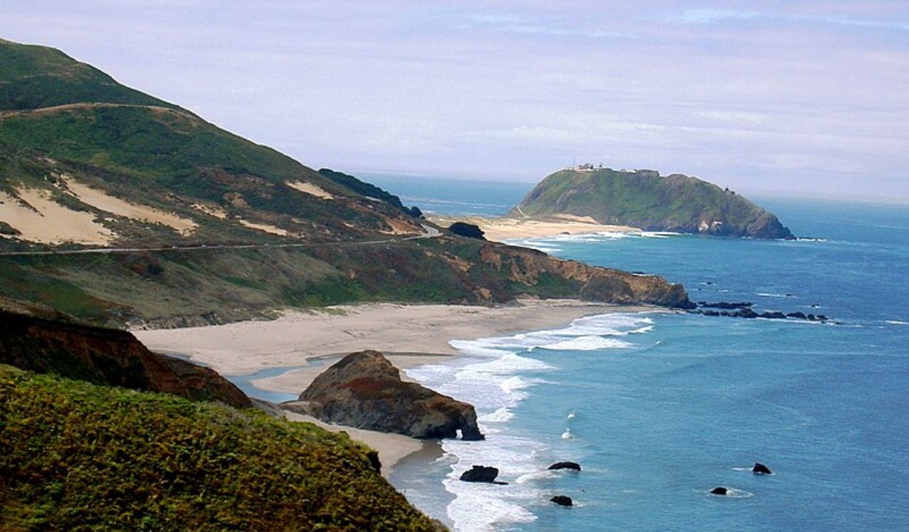 Rocky coastline with sandy beaches, green hills, and a large distant island beneath a partly cloudy sky.