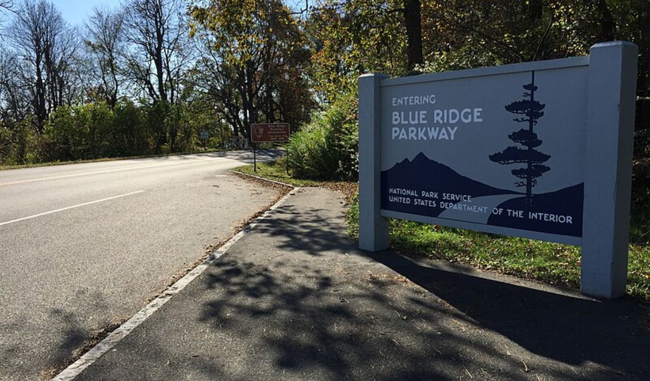 A large roadside sign reads "Entering Blue Ridge Parkway, National Park Service, United States Department of the Interior," with trees and a clear sky in the background.