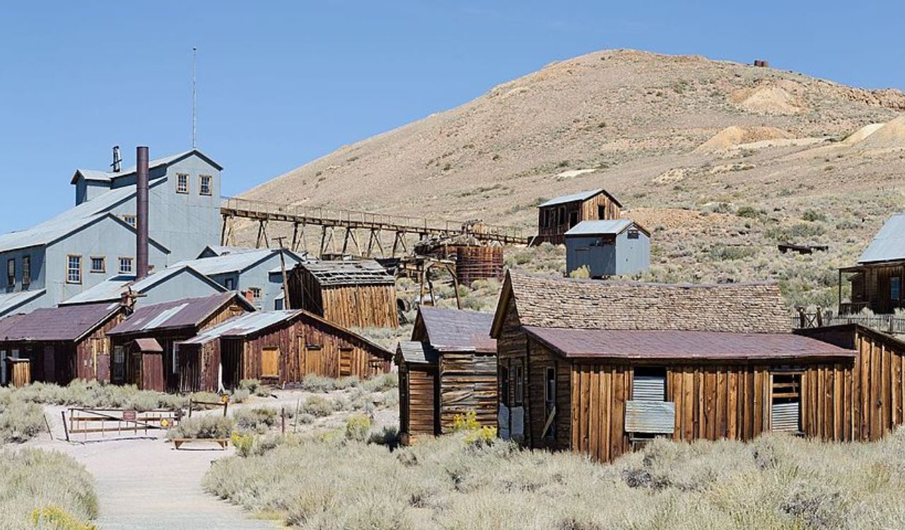 Old wooden and metal buildings of an abandoned mining town stand in a dry, scrubby landscape beneath a clear sky and a distant hill.