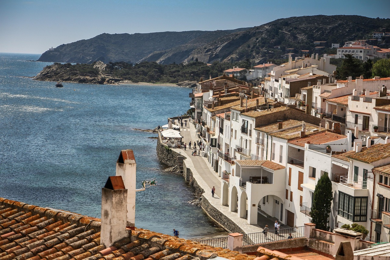 White buildings line a narrow seaside promenade in a coastal town, with hills and blue water beneath a clear sky.