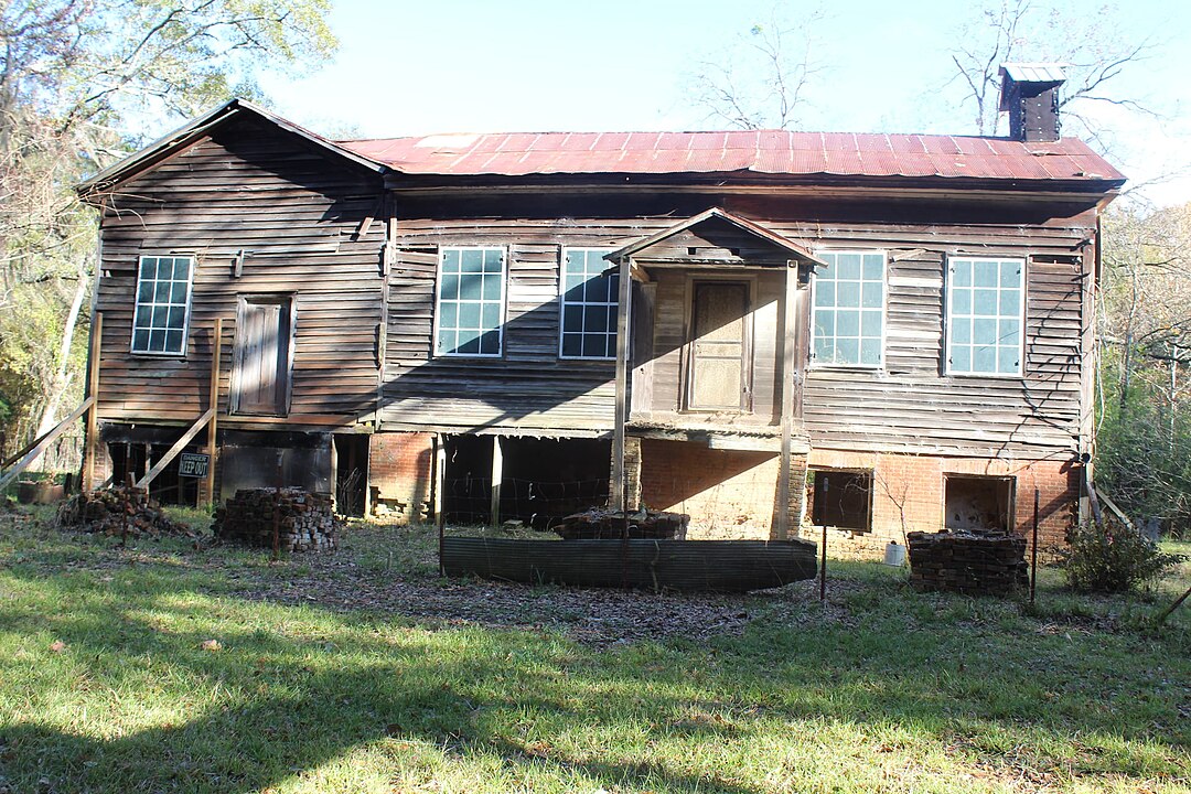 A weathered wooden house with missing planks, a rusty roof, and broken windows stands on brick supports in a grassy yard.