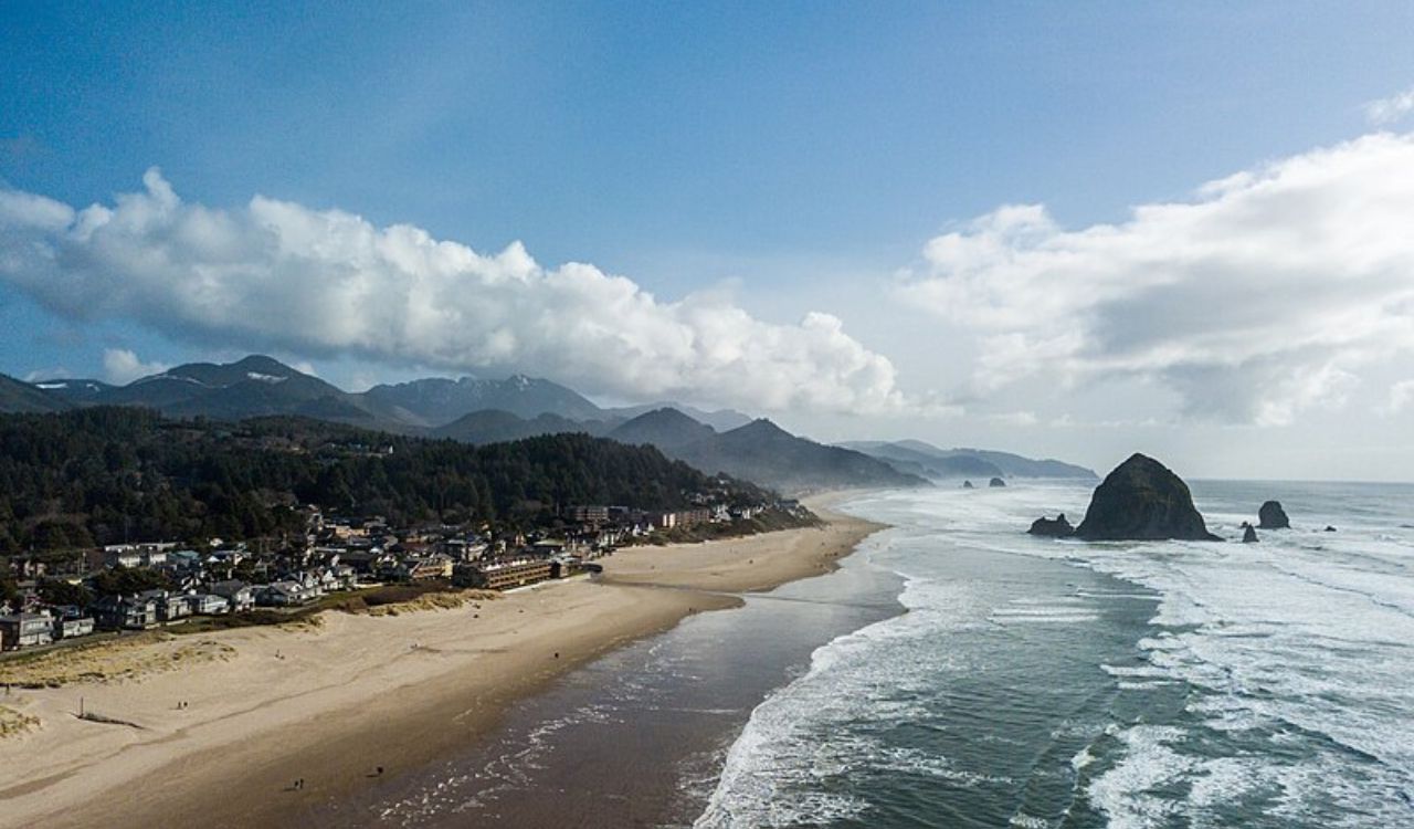 A wide sandy beach with gentle waves, large sea stacks, a coastal town, and tree-covered mountains sits under a partly cloudy sky.