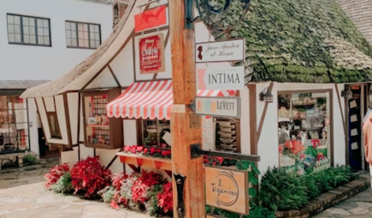 A small, quaint shop with a moss-covered roof, red-and-white striped awning, and flower displays stands on a cobblestone street.