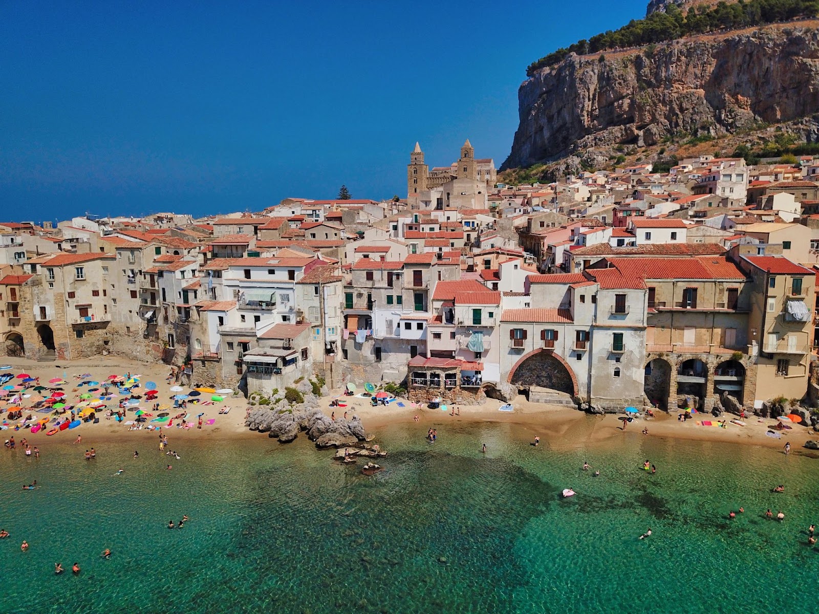 A coastal town with historic red-roofed buildings borders a turquoise sea, where crowds swim and sunbathe on the beach.