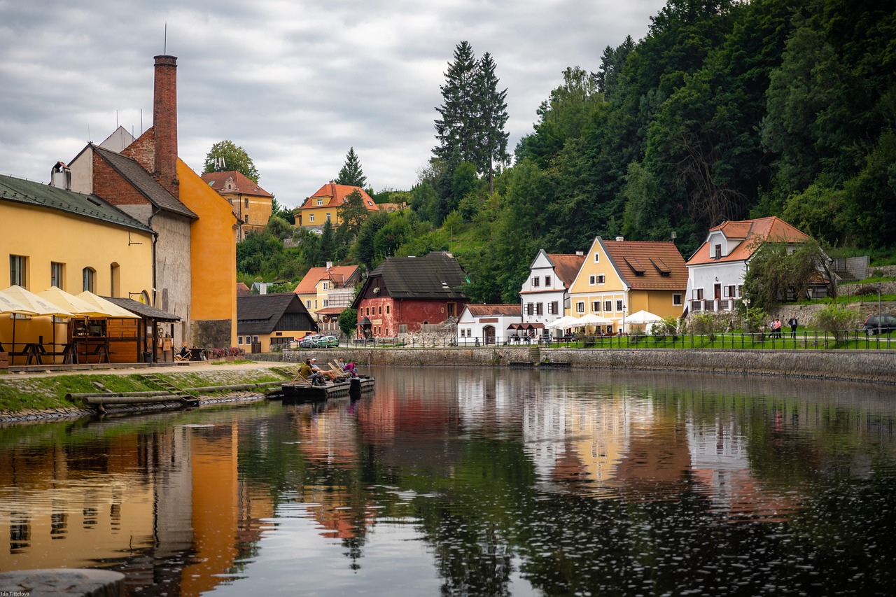 A small riverside village with colorful houses, a tall chimney, outdoor seating, and lush trees is reflected in the water beneath a cloudy sky.