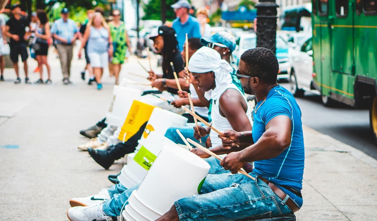 Street performers play white plastic buckets as drums on a sidewalk, with a green vehicle and pedestrians in the background.