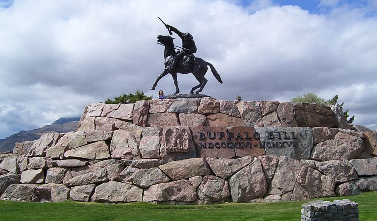 A bronze equestrian statue inscribed "Buffalo Bill" with Roman numerals stands atop a large stone monument outdoors amid grass and trees.