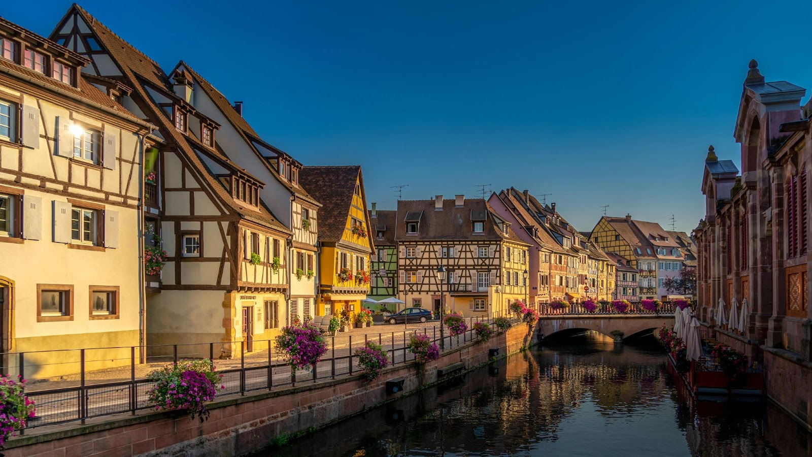 Half-timbered houses with flower boxes line a canal spanned by a stone bridge in a European town by day.