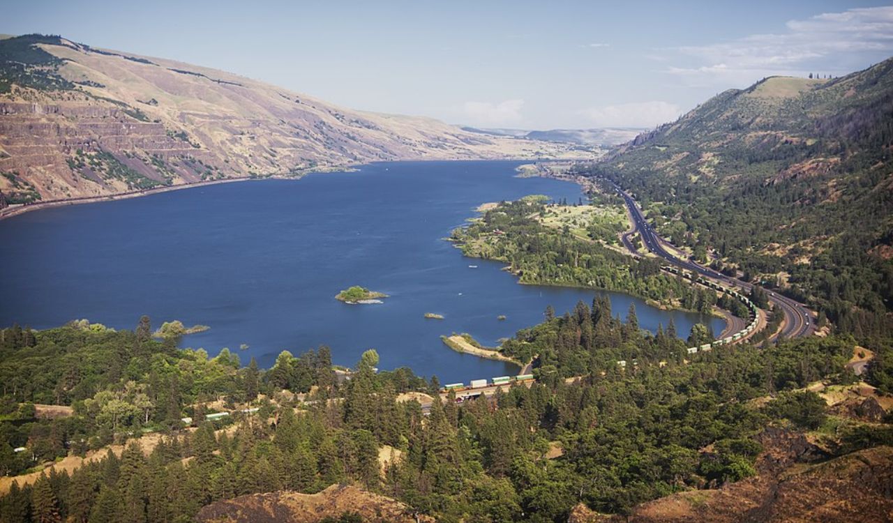 A winding river flows through a green valley bordered by forests, a highway, and mountains beneath a clear blue sky.