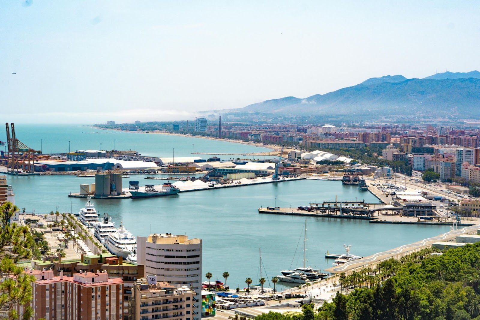 Aerial view of a coastal city featuring a marina with yachts, an industrial port, buildings, and mountains in the background.