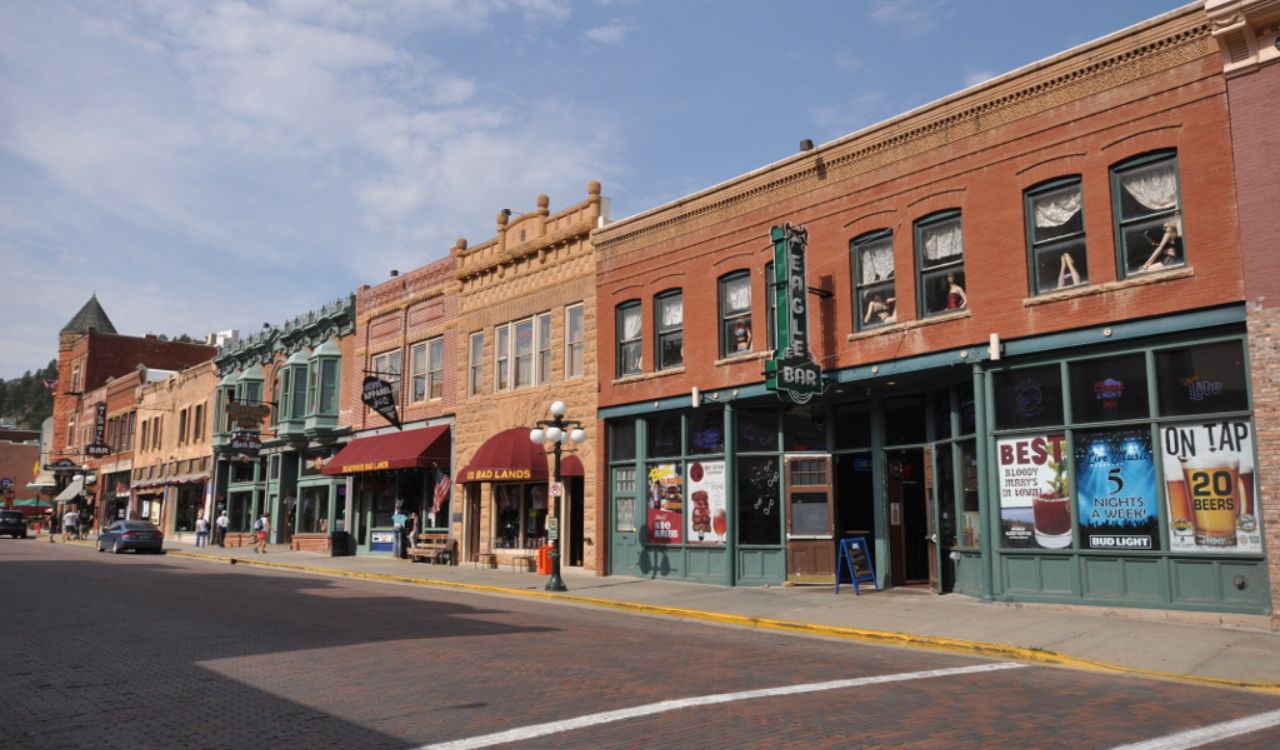 Historic brick buildings with bars, restaurants, and shops line a quiet, empty street beneath a partly cloudy sky.