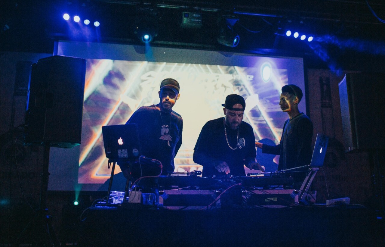 Three DJs perform on stage with electronic equipment and laptops, lit by blue lights and a colorful screen.