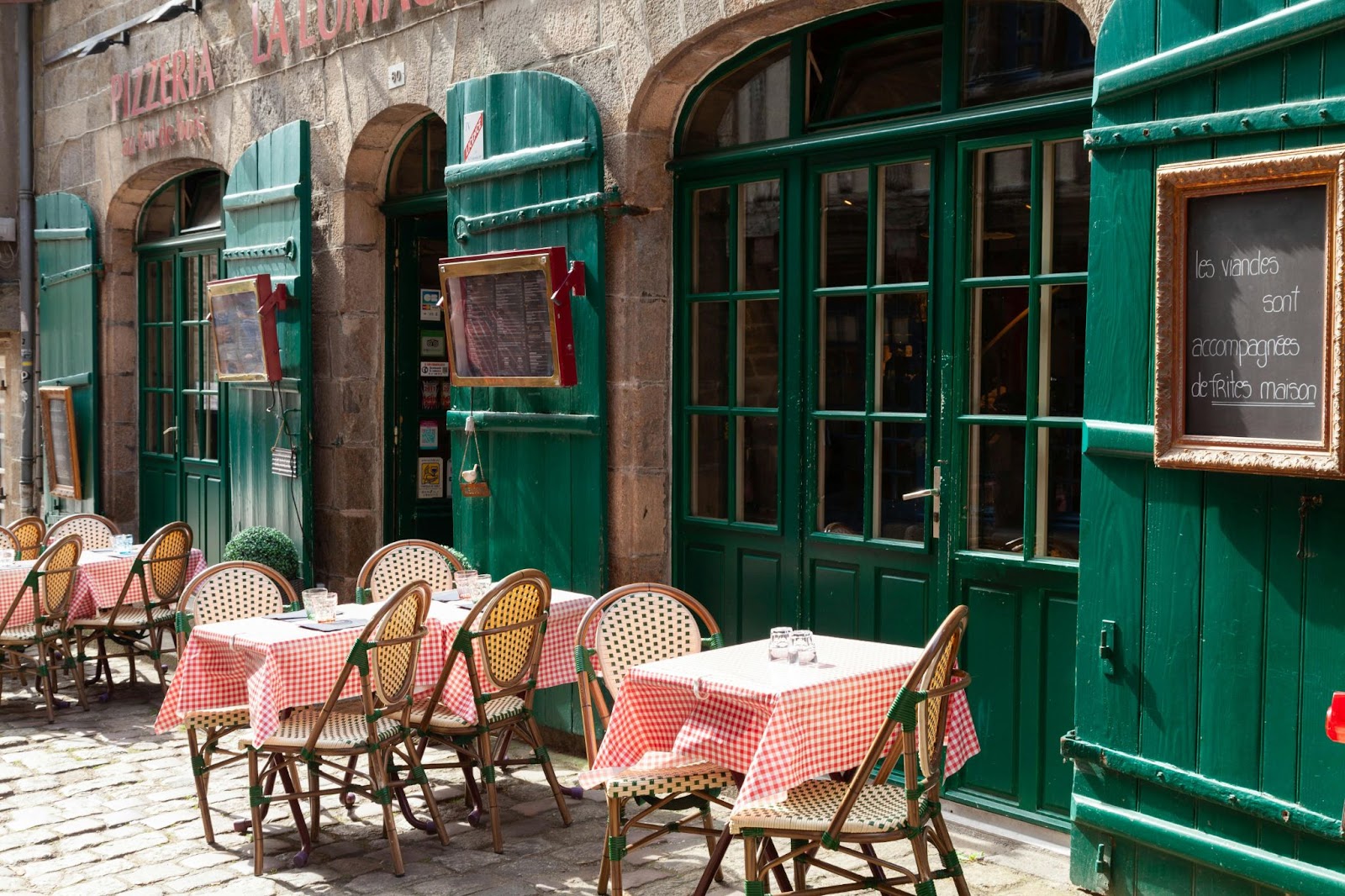 Outdoor tables with red checkered cloths and chairs sit before a restaurant with green doors and windows on a cobblestone street.
