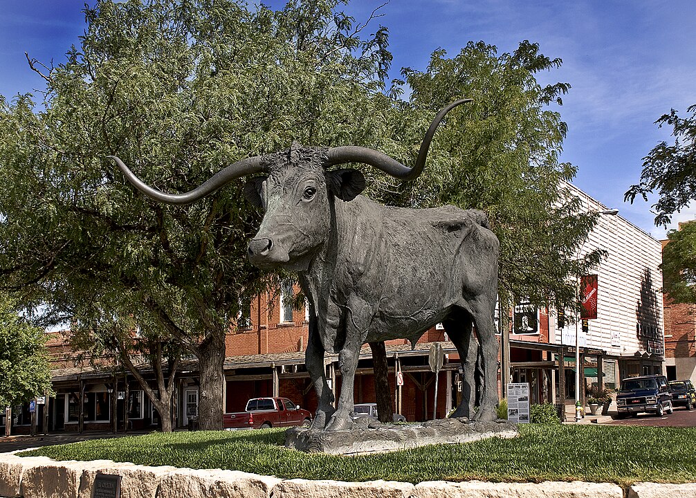 A bronze longhorn steer statue stands on a stone pedestal amid trees and storefronts in a small town.