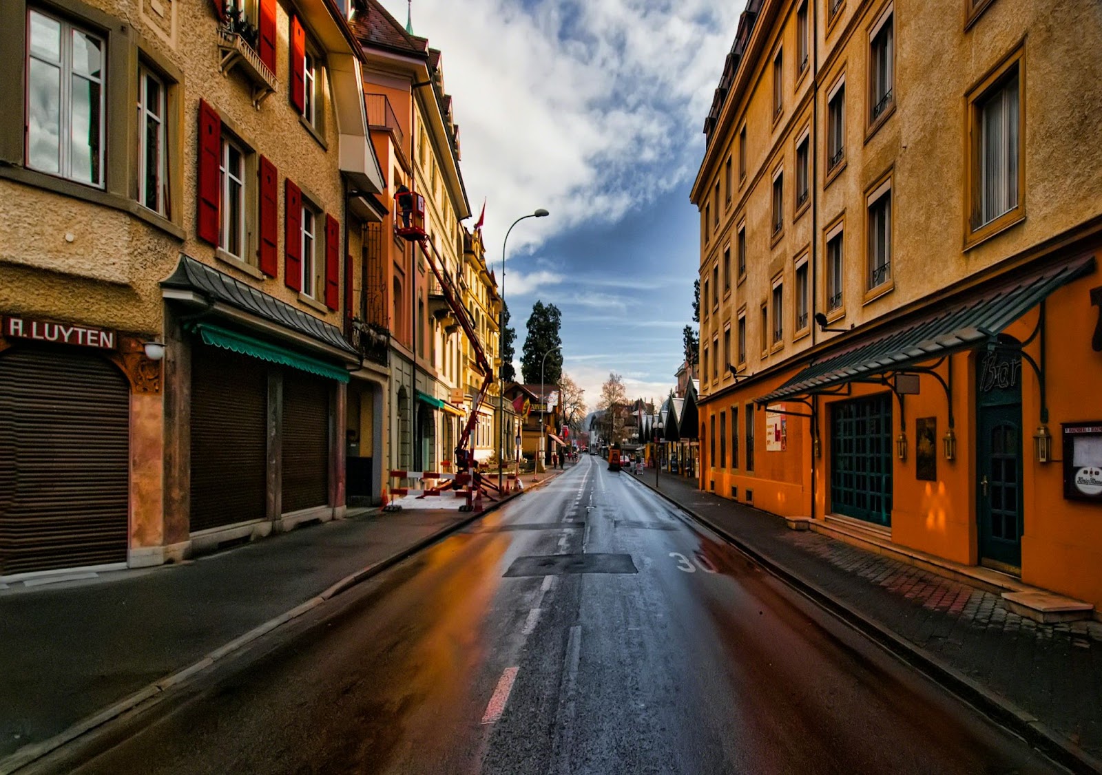 A wet, empty street divides colorful, tightly packed buildings beneath a partly cloudy sky in a small town.