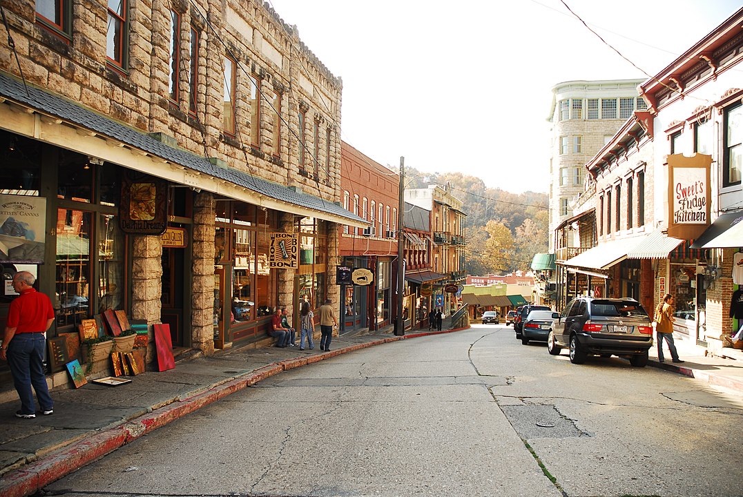 A narrow, sloped street in a small town is lined with historic brick and stone buildings, shops, parked cars, and a few pedestrians.