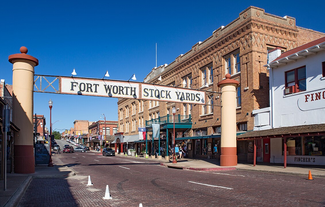 The Fort Worth Stock Yards entrance sign spans a brick road lined with historic buildings and shops under a clear blue sky.