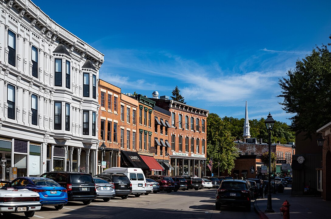 A downtown street features parked cars, historic brick and white buildings, storefronts, and a church steeple beneath a blue sky.