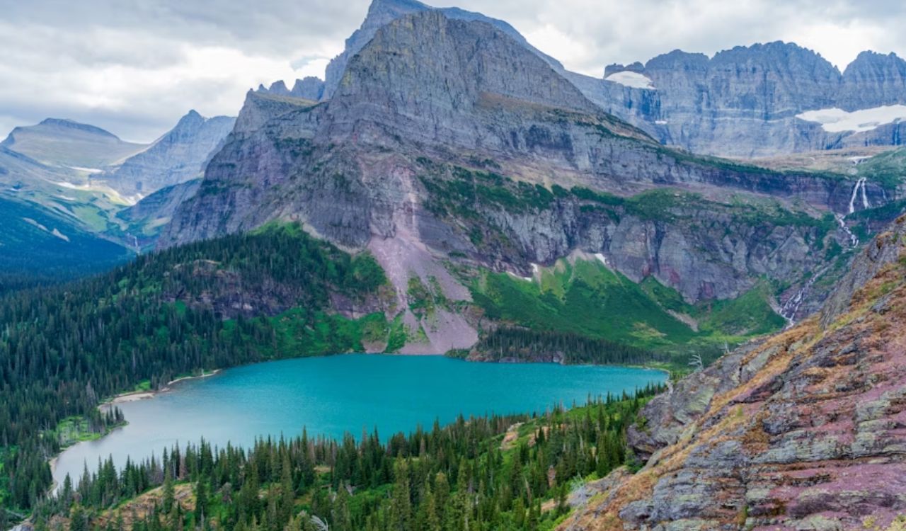 A turquoise alpine lake is surrounded by dense forest and rugged mountain peaks beneath a cloudy sky.