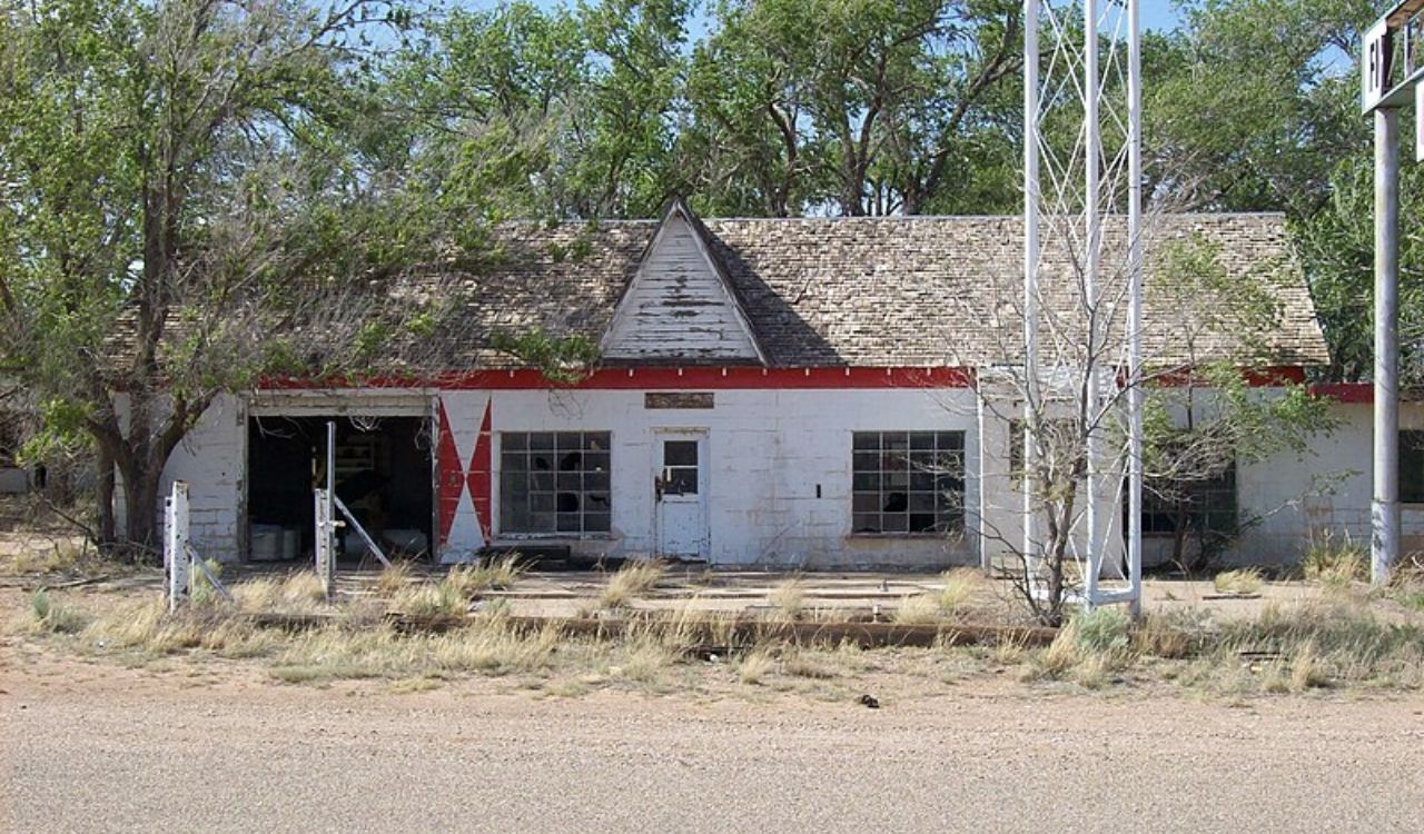 An abandoned, white-painted building with boarded windows stands beside a road amid overgrown grass and trees.