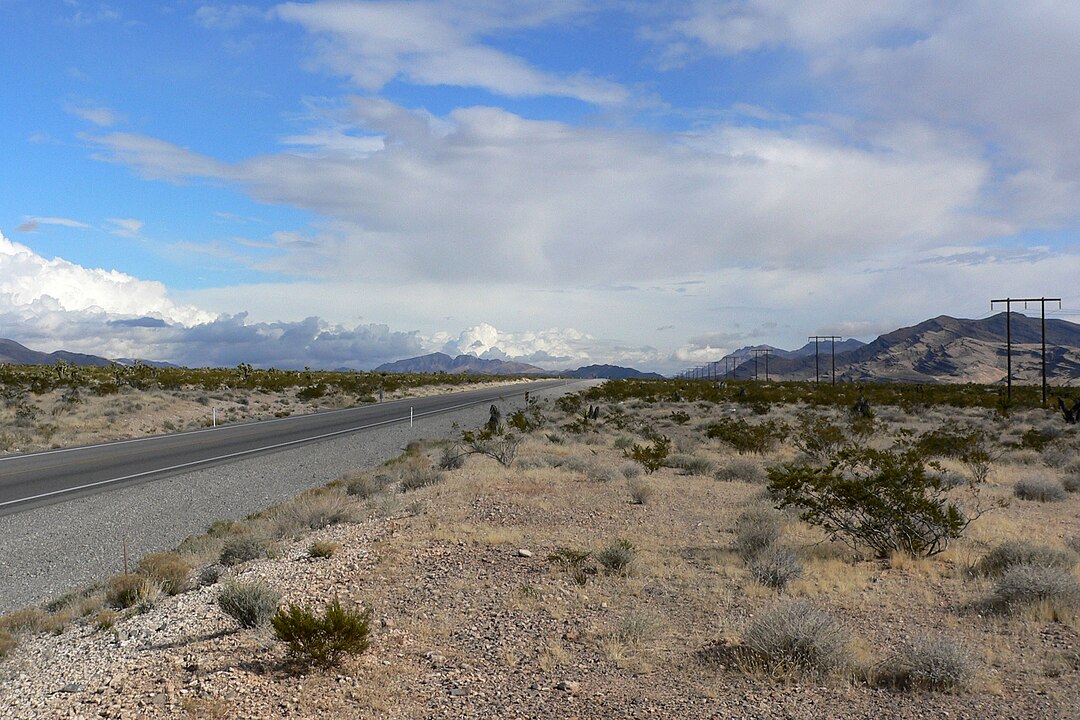 A paved road cuts through a dry desert with sparse vegetation, distant mountains, and a partly cloudy sky.