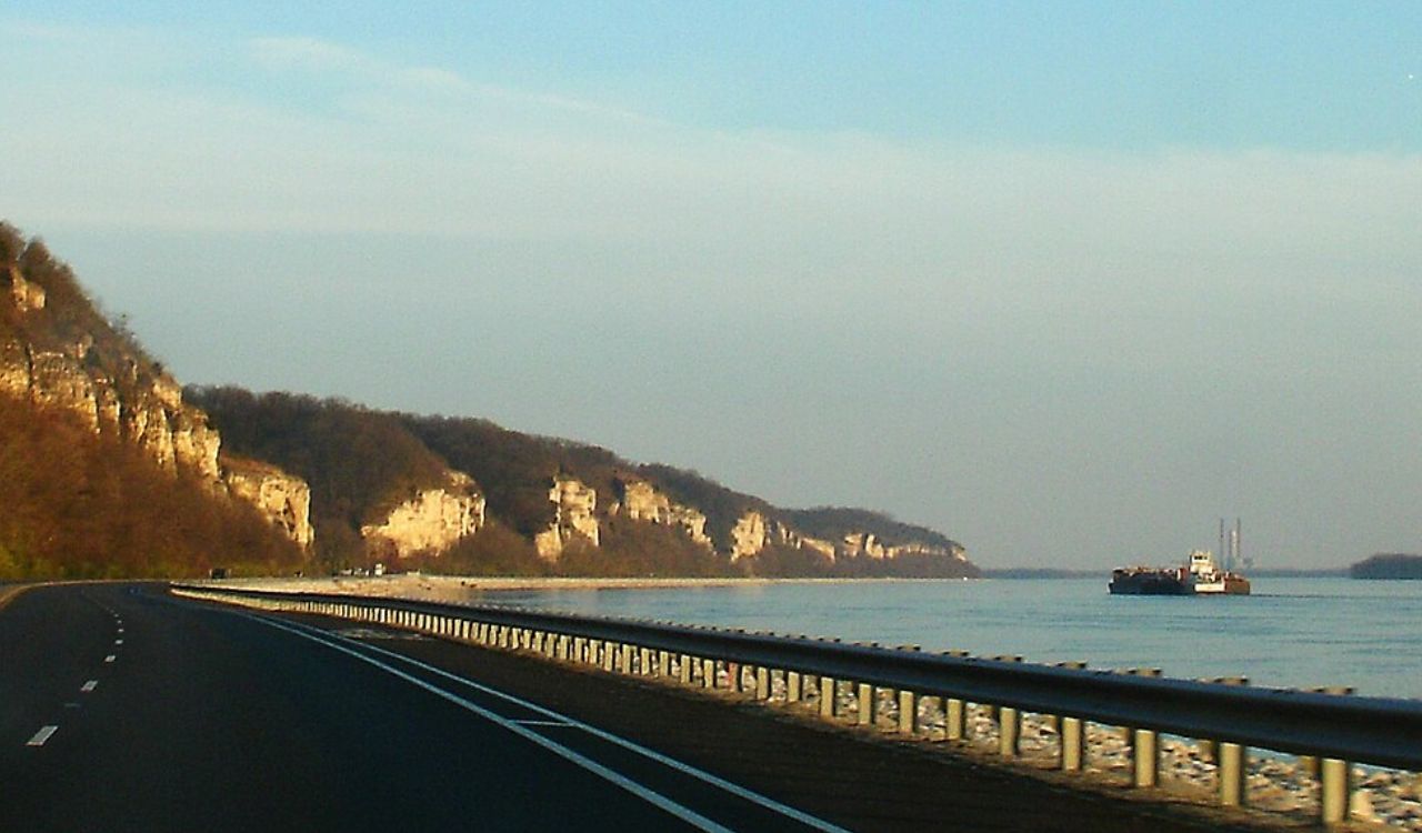 A paved road with a metal guardrail runs beside a river, bordered by forested cliffs on the left and a barge on the water to the right under a clear sky.