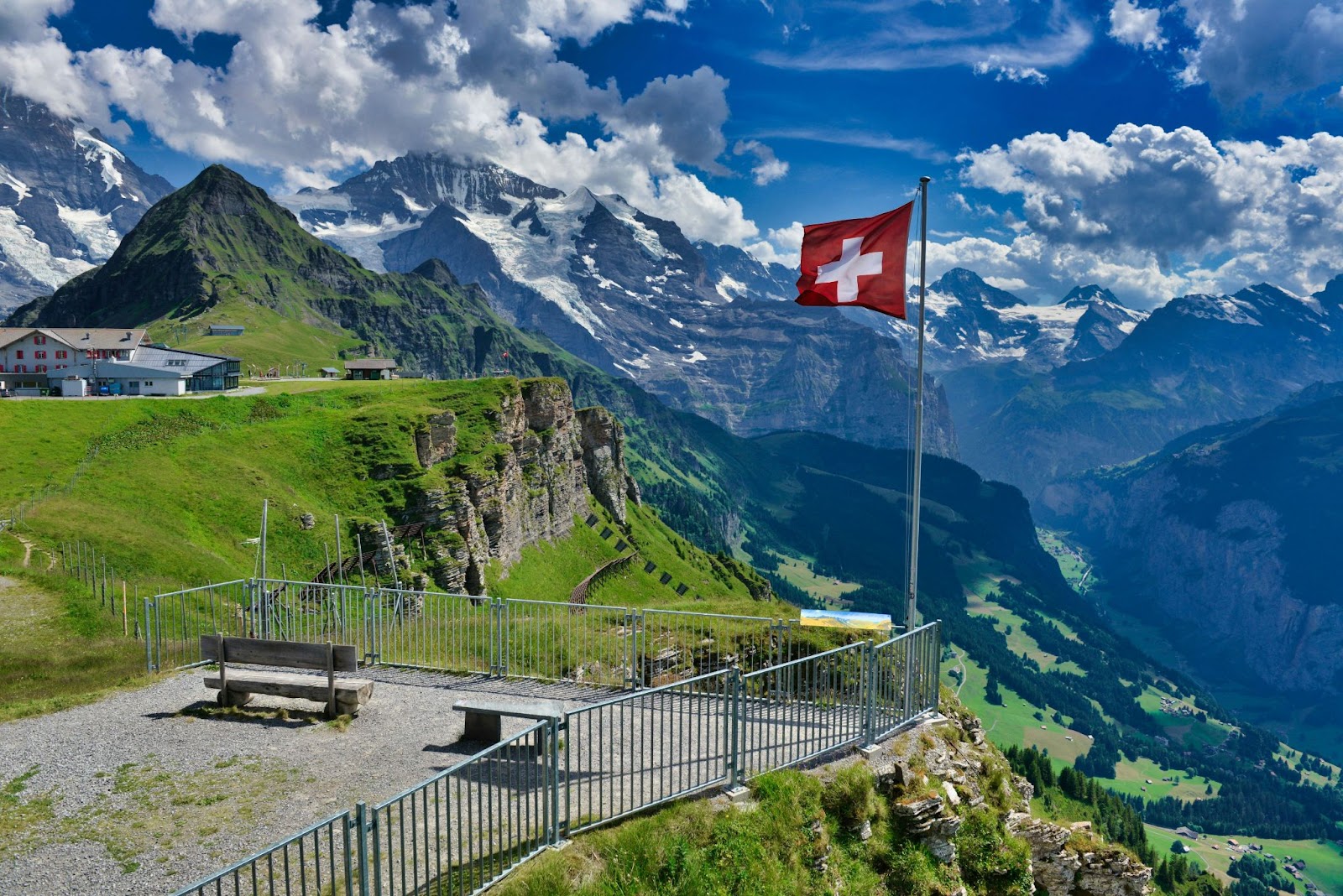 A Swiss flag waves beside a bench overlooking a green valley and snow-capped mountains under a partly cloudy sky.