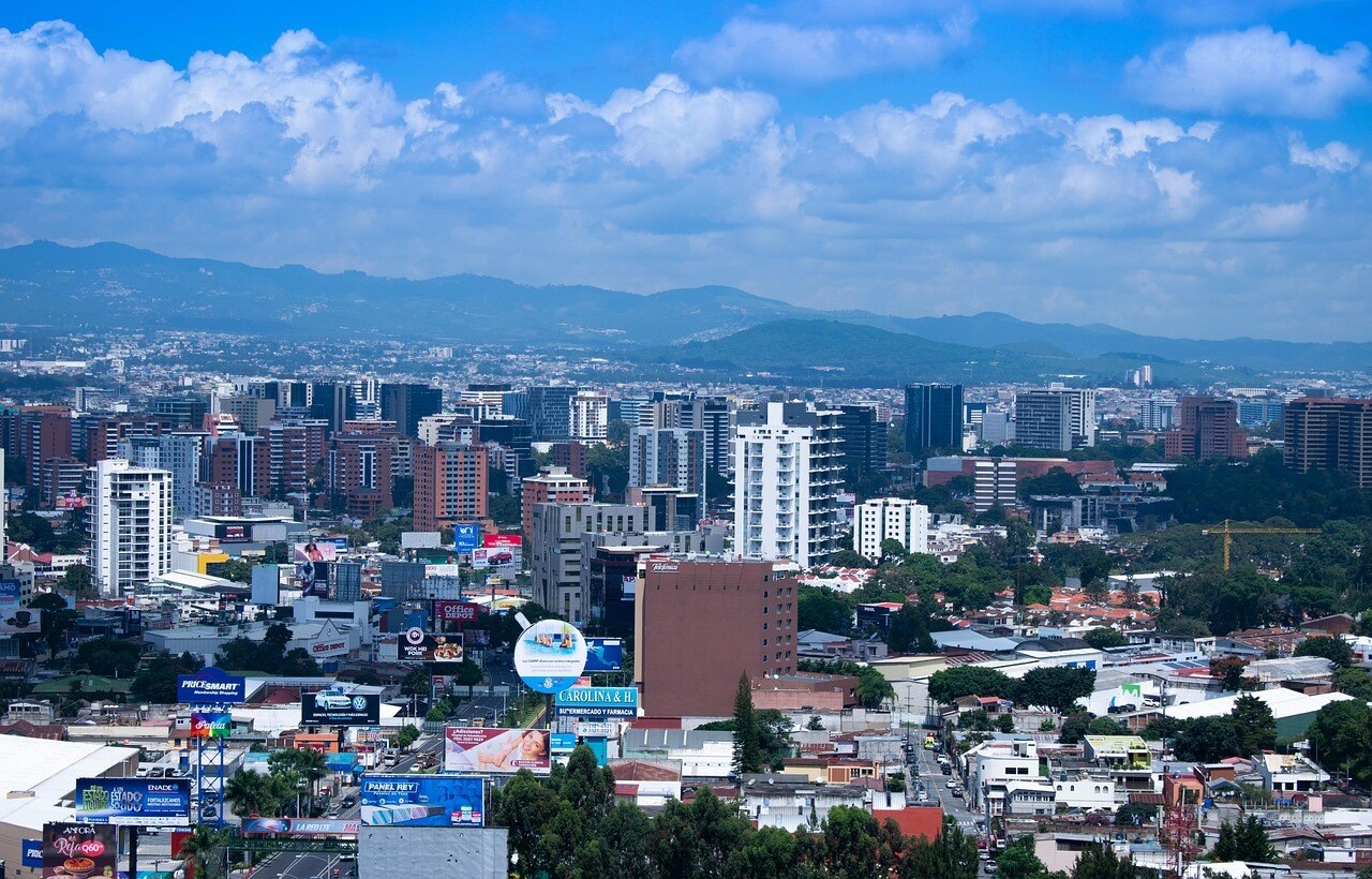 A cityscape features modern buildings, billboards, and apartments beneath a blue sky with scattered clouds and mountains in the background.