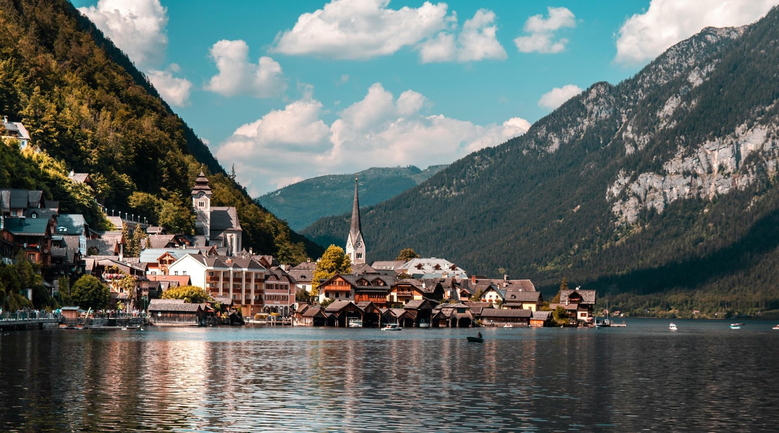An alpine-style lakeside village with a tall-steepled church sits amid forested mountains beneath a partly cloudy sky.