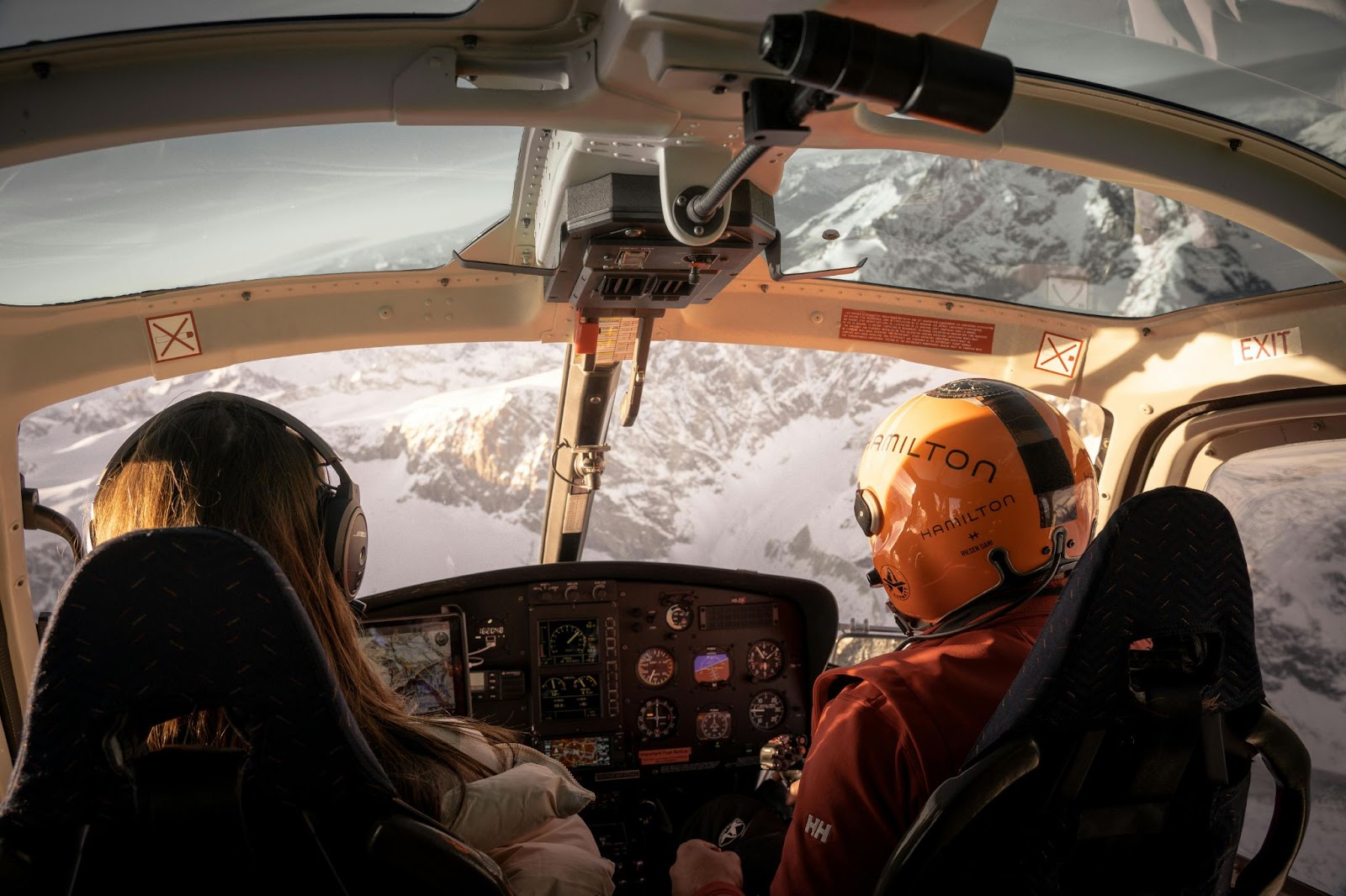 Two people wearing headsets sit in a helicopter cockpit flying over snowy mountains, with visible instrument panels and controls.