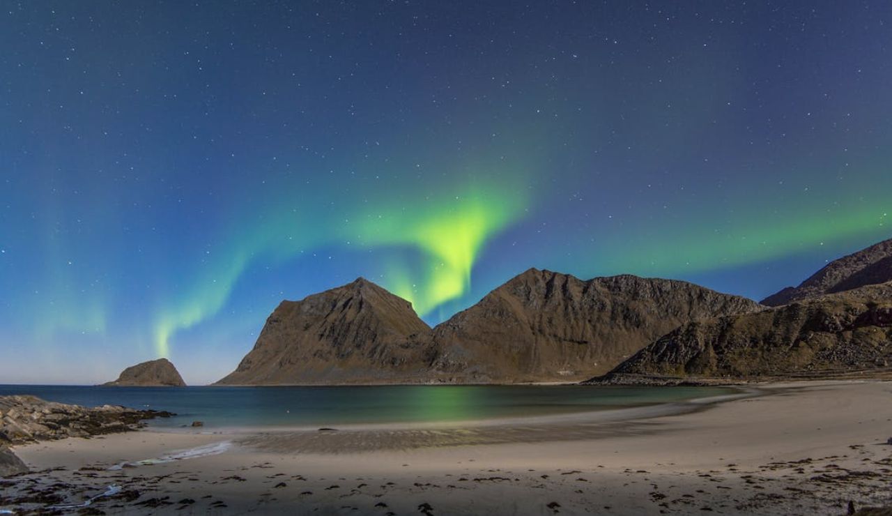 Northern lights glowing above rugged mountains and a sandy shoreline in Hemnes, Norway.