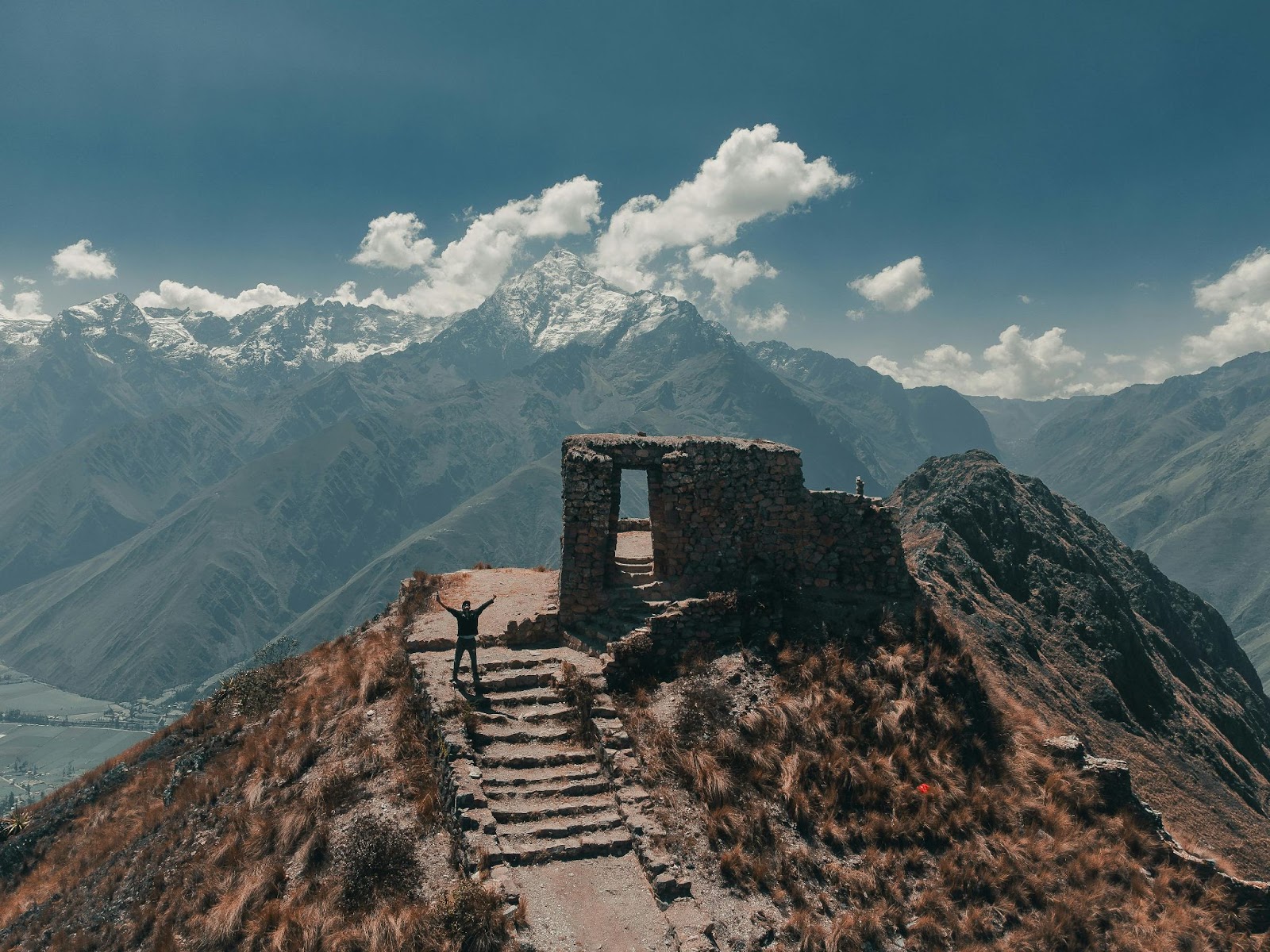 A person stands by ancient stone ruins atop a mountain overlooking snow-capped peaks and a valley under a partly cloudy sky.