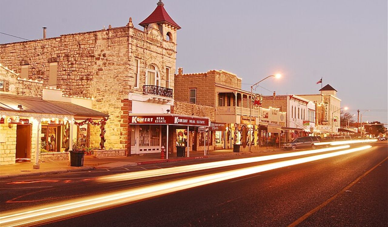 At dusk, light trails from cars illuminate a small-town main street with historic stone buildings.