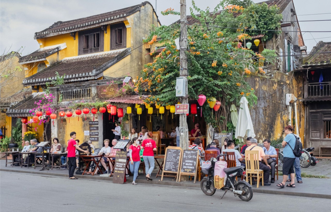 People sit and walk outside a busy yellow-walled cafe adorned with lanterns, menus, and plants.