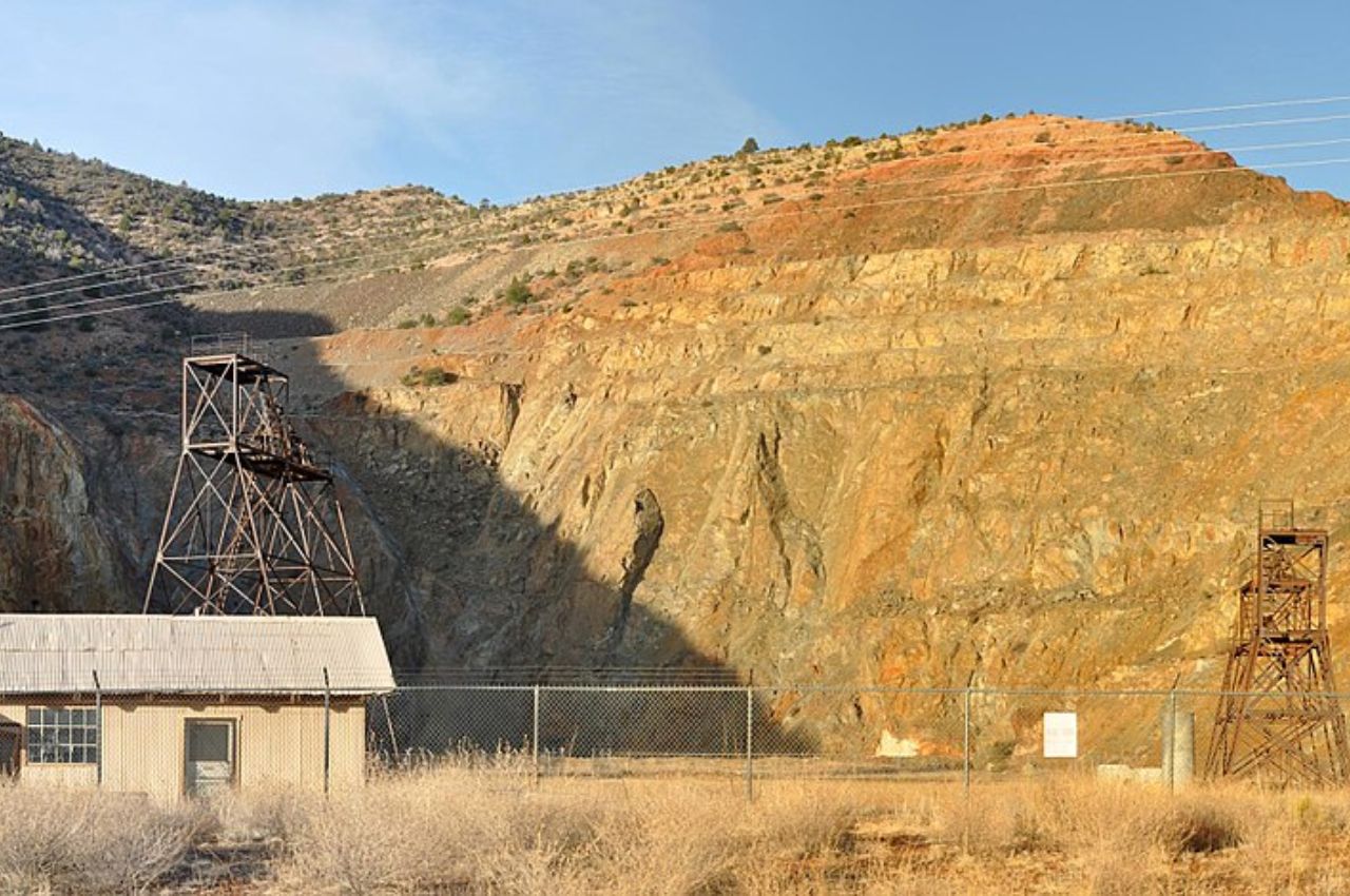 An old mining site with rusted metal structures, a small building, and a fence sits before a rocky hillside under a clear sky.