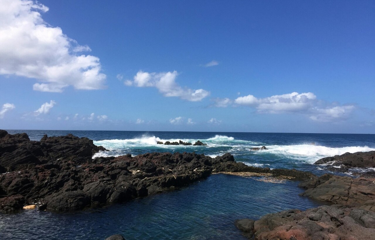 Waves crash against a rocky shoreline of black volcanic rocks and a small tidal pool beneath a blue sky with scattered clouds.