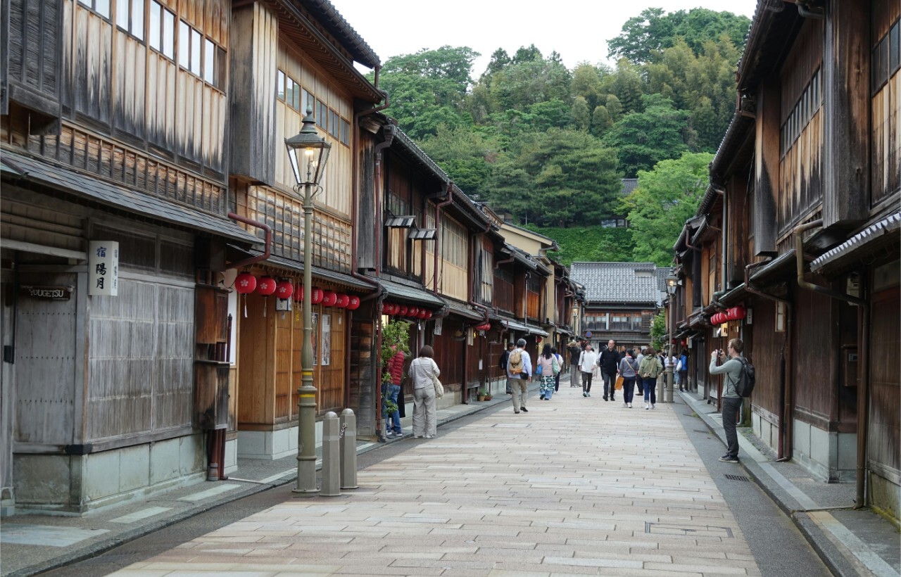A stone-paved street lined with traditional wooden buildings and red lanterns, with people walking and greenery in the background.