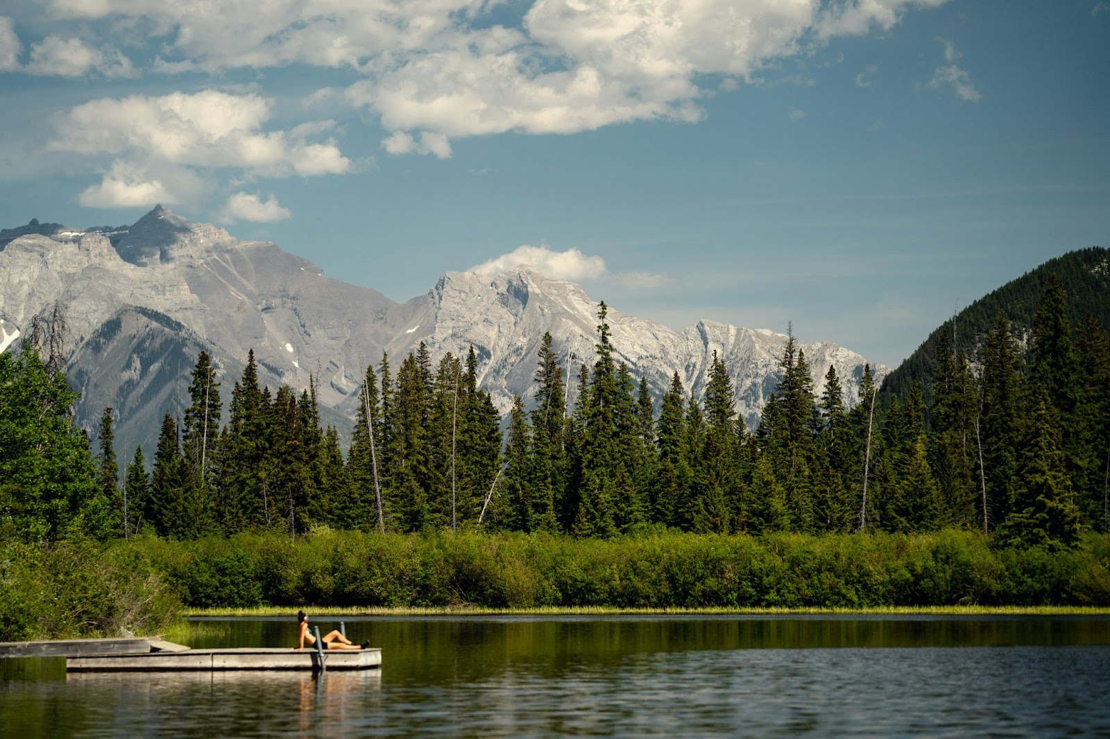 Two people sit on a small wooden dock by a calm lake, surrounded by evergreen trees and mountains beneath a partly cloudy sky.