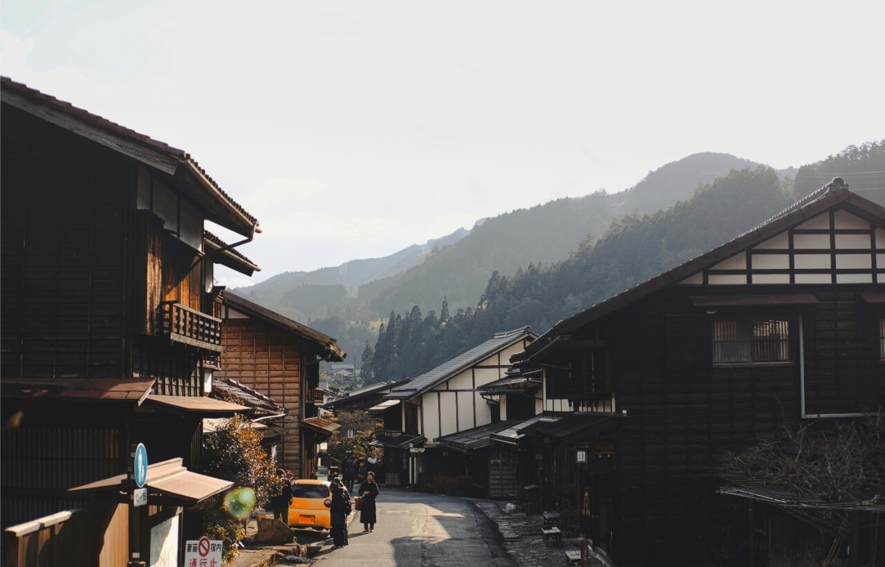 A quiet Japanese village street features wooden houses, a parked yellow car, and mountains beneath a hazy sky.