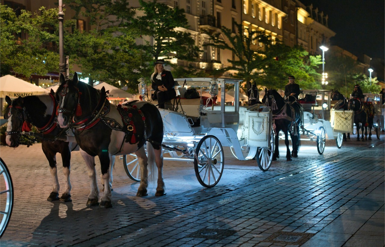At night, horse-drawn carriages with lit lamps and drivers sit along a cobblestone street, with buildings' illuminated windows in the background.