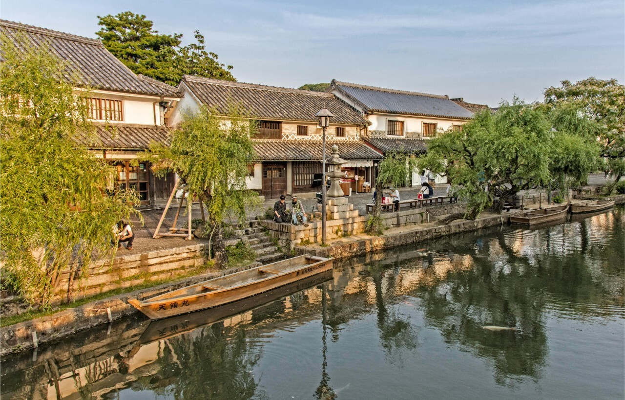 Traditional Japanese buildings line a calm canal with small boats, steps to the water, trees, and a few people along the walkway.