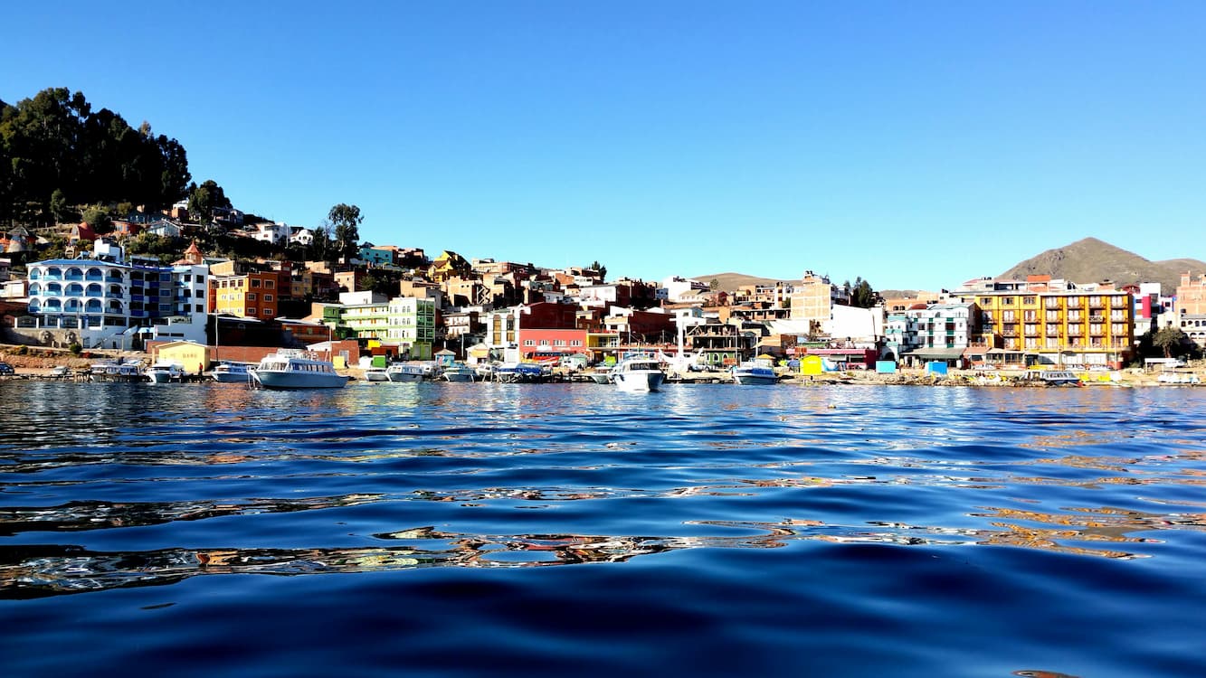 Colorful buildings line a waterfront beneath a clear blue sky, with boats floating on calm, reflective water in the foreground.