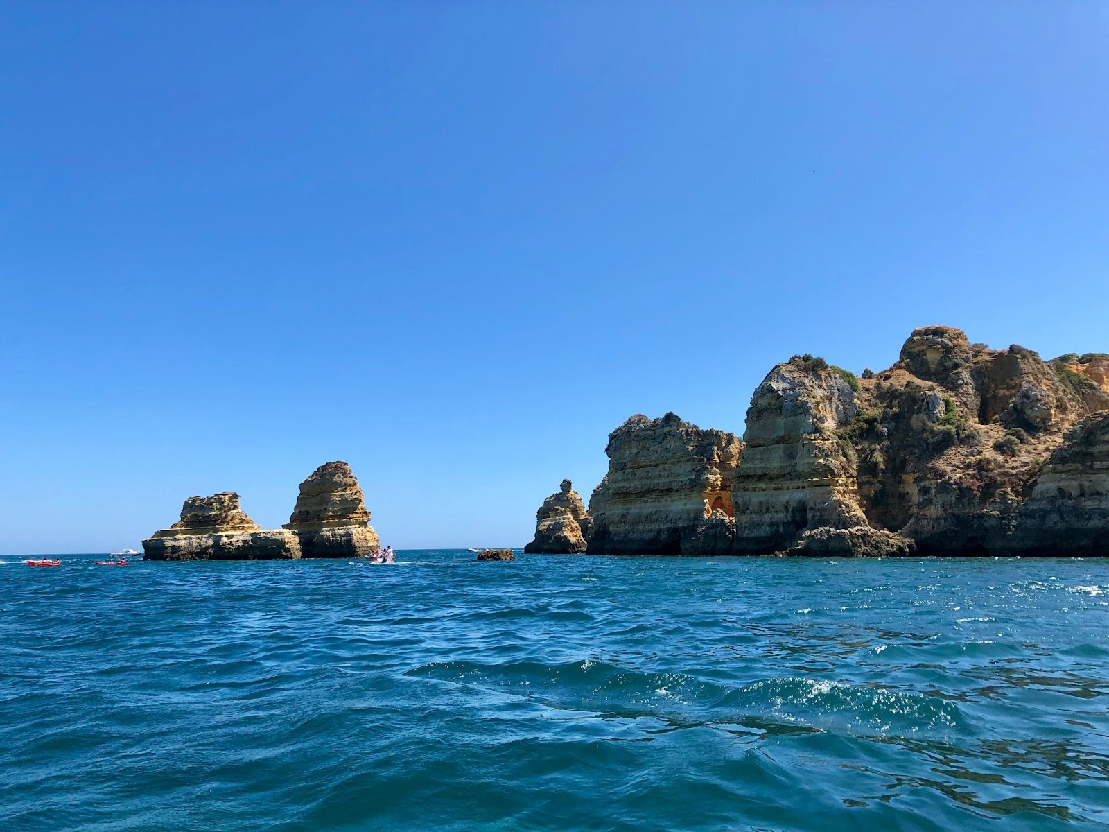 Rocky cliffs and sea stacks rise from blue water beneath a clear sky, with calm waves in the foreground.