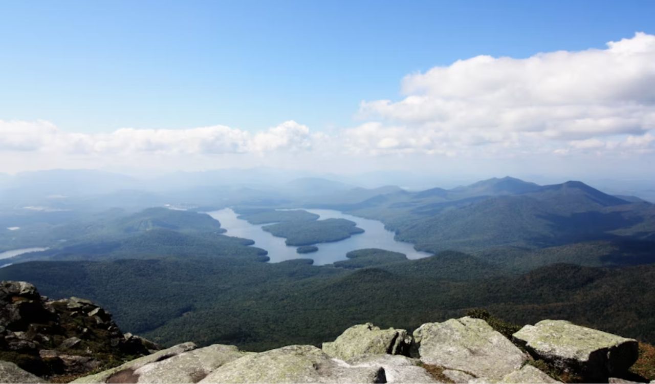 A rocky mountaintop overlooks a winding lake encircled by dense forest and distant hills beneath a partly cloudy sky.
