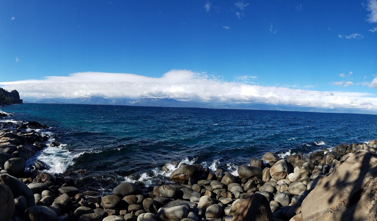 Waves crash on rocky shoreline beside deep blue water and distant mountains beneath a bright, cloud-scattered sky.