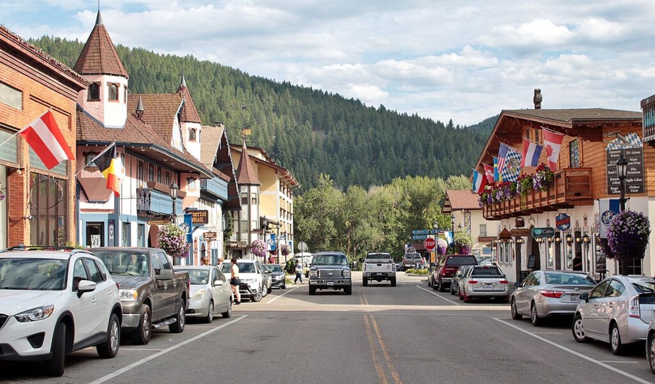A small town main street with parked cars, alpine-style buildings, and national flags sits before forested hills.