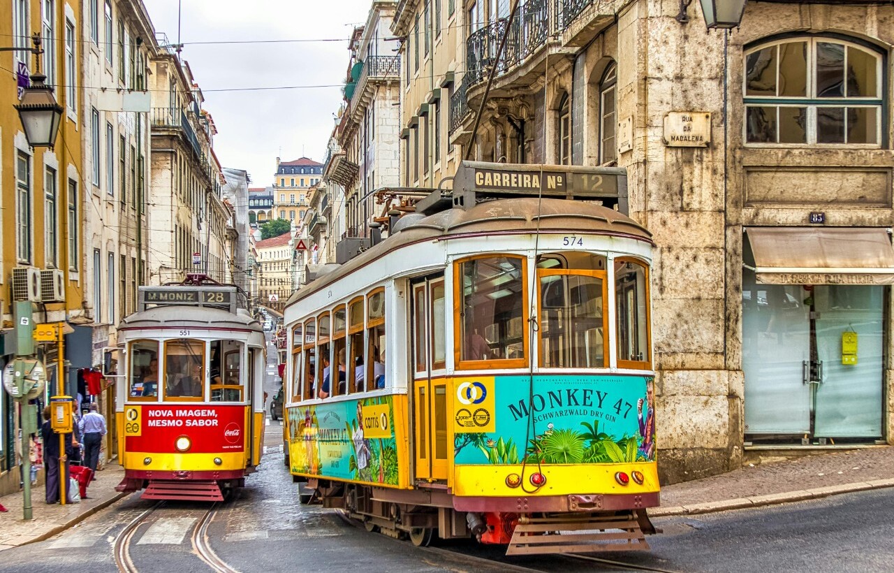 Two vintage yellow trams pass through a narrow street of old buildings in Lisbon, Portugal.