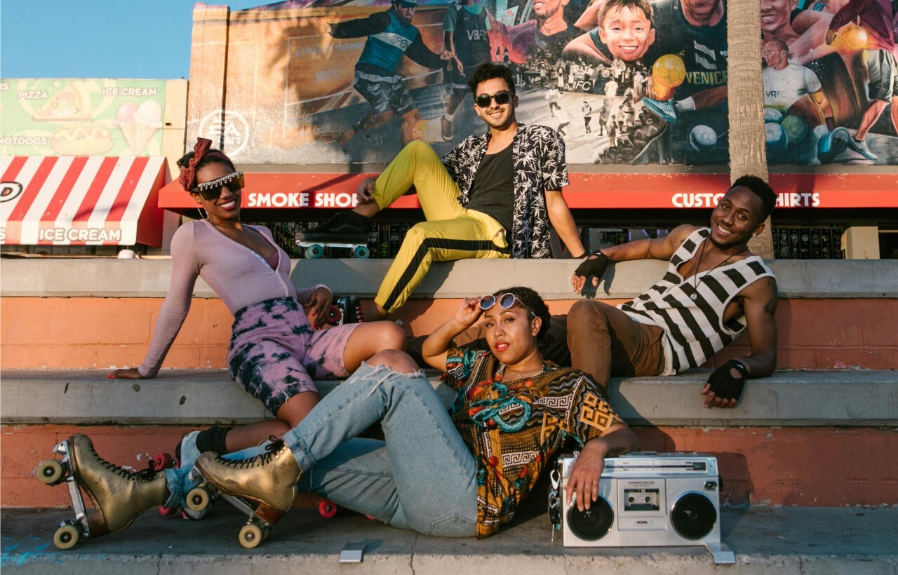 Four young adults pose on outdoor steps, two in roller skates and one holding a boombox, with a colorful mural and storefronts behind them.
